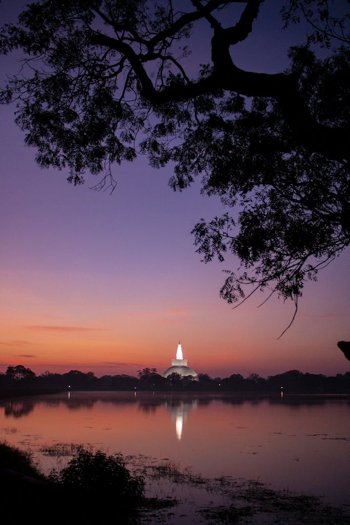 A tranquil sunset scene with a pagoda reflecting on a calm lake, framed by silhouetted trees.