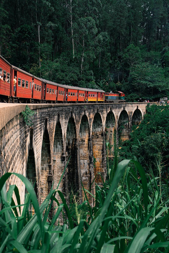 A vibrant red train travels across the historic Nine Arches Bridge in Ella, Sri Lanka, surrounded by lush greenery.