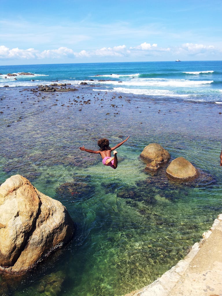 Person joyfully jumping into the clear ocean waters by the rocky Sri Lankan shore, embracing summer thrills.